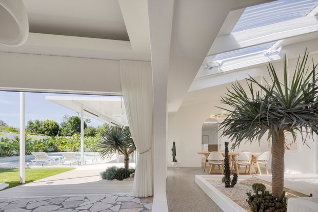 Bright, airy contemporary interior opening onto a landscaped outdoor area and pool. The space features a polished light floor, white walls and curtains, large glazed openings, a dramatic slatted skylight, and a raised interior planter filled with sculptural desert plants. A dining area is visible beyond the planter, while the adjacent covered terrace has pale seating and patterned stone paving.