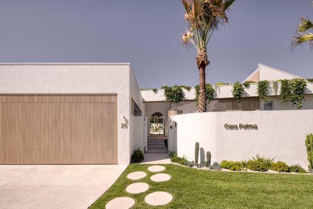 Front exterior of a low-rise contemporary residence with white rendered walls, a large timber-look slatted garage door, gated entry, landscaped lawn, circular stepping stones, cacti, palms, and climbing greenery. The flat-roofed facade has clean geometric forms and a warm, resort-like material palette.