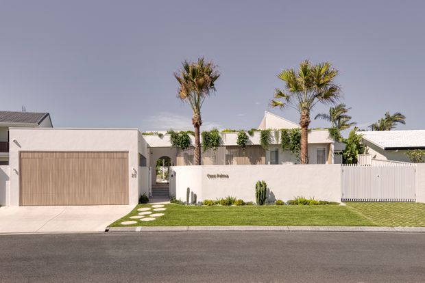 Street-facing view of a contemporary single-storey residential property with a pale rendered facade, attached garage, white gated entry, flat and low-pitched roof forms, and a manicured front lawn. Tall palms, sculptural stepping stones, and sparse planting create a clean resort-like frontage.