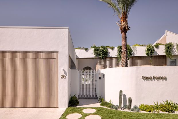 Street-facing exterior of a contemporary residence named Casa Palma. The white rendered facade features a large vertical timber-slatted garage door, matching slatted screening, a gated entry, minimalist landscaping with cacti and a palm tree, and a low flat roof with a small pitched-roof section visible at right.