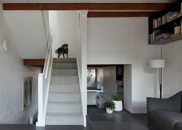Compact contemporary living area with a carpeted staircase, white-painted masonry walls, exposed dark timber ceiling beams, dark tiled flooring, and built-in black shelving. A partial kitchen is visible through the opening beneath the stairs, while a grey sofa and floor lamp occupy the foreground.