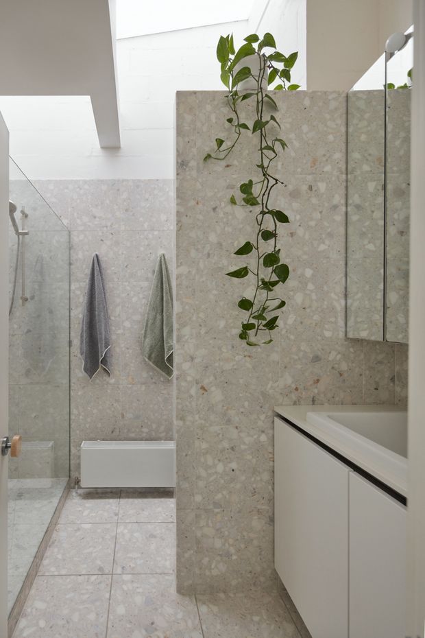 Bright contemporary bathroom featuring pale terrazzo-style wall and floor tiles, a frameless glass shower enclosure, wall-mounted white toilet, floating white vanity, large mirror, and a hanging trailing plant. A rooflight or clerestory-like opening provides abundant natural light.