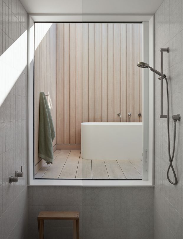 A bright contemporary bathroom viewed through a large interior window or glazed partition. The foreground contains a walk-in shower with grey tiled walls, exposed chrome shower hardware and a small timber stool. Beyond the glazing is a freestanding white bathtub set against a vertical light timber-panelled wall, with pale timber-look flooring and a hanging towel. Strong daylight creates a clean, calm, refined atmosphere.