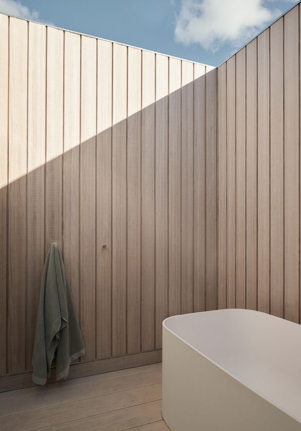 A minimalist open-air or enclosed bathing space featuring a freestanding white bathtub in the foreground, vertical natural timber wall panelling, and matching timber floorboards. Strong sunlight creates a diagonal shadow across the wall, with a muted towel hanging from a simple hook. A small opening at the top reveals blue sky and clouds, contributing to an airy, serene atmosphere.