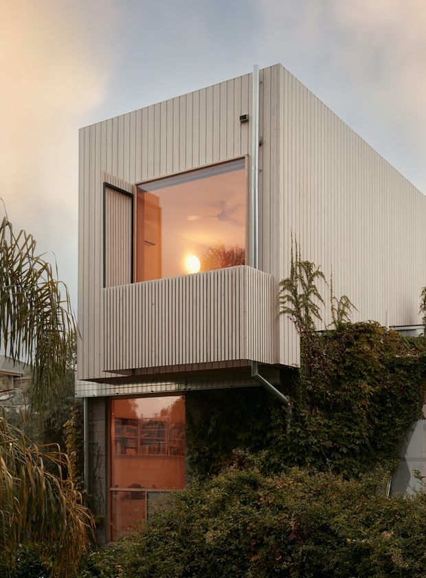 Exterior view of a compact contemporary multi-storey house with a strongly geometric rectangular form. The facade is clad in pale vertically ribbed weatherboard-like panels, with warm-toned glazed openings, a projecting upper balcony enclosed by matching vertical slats, exposed downpipes, and dense climbing greenery around the lower level. The image is photographed at dusk, with warm interior light visible through the windows.