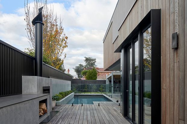 A contemporary residential courtyard viewed from a timber deck, featuring a rectangular swimming pool enclosed by frameless glass balustrades, a concrete outdoor fireplace with tall black flue, dark vertical fencing, and a timber-clad building with large black-framed glazed doors. The space has a restrained modern material palette and overlooks neighbouring residential rooftops and trees.