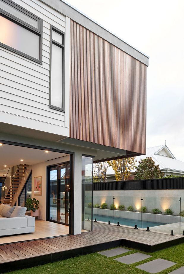 Contemporary two-storey house exterior with a projecting upper volume clad in narrow vertical timber battens, white horizontal weatherboard siding, dark window frames, and extensive ground-floor glazing. The open living room connects directly to a timber deck and a long narrow swimming pool in a landscaped rear courtyard.
