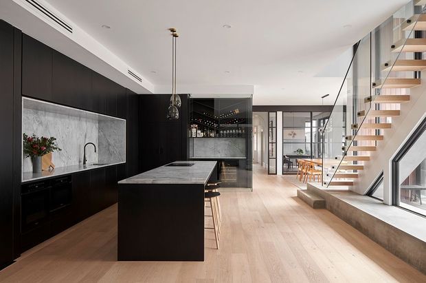 A contemporary open-plan kitchen and dining area with a dramatic dark timber kitchen, large marble-look splashback and island, integrated appliances, pendant lighting, and timber bar stools. Pale timber flooring extends toward a light-filled dining space, while a floating timber staircase with glass balustrades and expansive glazing defines the right side of the frame.
