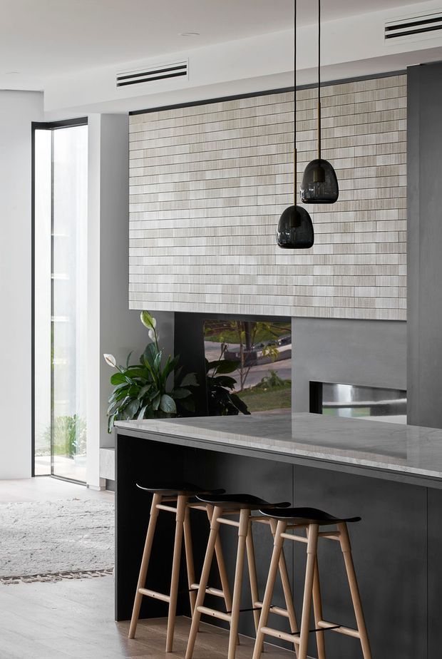 A refined contemporary kitchen with a long dark island and integrated seating, topped by a pale stone benchtop. The island faces a textured tiled feature wall with a built-in fireplace and a dark reflective opening. Two black pendant lights hang above the island, while full-height glazing at left brings in natural light and views of greenery. The palette is predominantly charcoal, grey, pale stone, and natural timber.