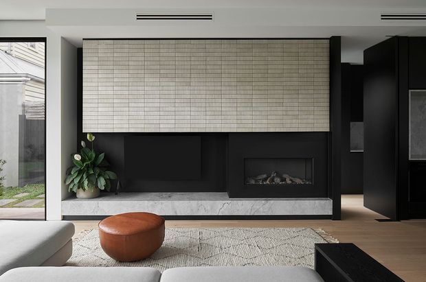 Contemporary living room with a dramatic black feature wall incorporating a built-in fireplace and large textured tiled panel. A pale stone hearth, light timber flooring, neutral rug, curved light sofa, and round brown leather ottoman create a refined, softly contrasting interior. Floor-to-ceiling glazing at left opens toward a small landscaped courtyard, while dark built-in cabinetry is visible at right.