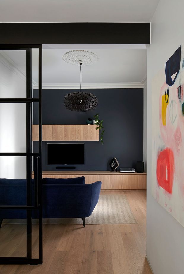 A refined living room viewed through a black-framed glazed room divider. The space features a dark navy feature wall, light timber built-in television cabinetry and low storage, a deep blue sofa, pale timber flooring, ornate ceiling cornice and ceiling rose, a perforated dark pendant light, and colourful abstract wall art.