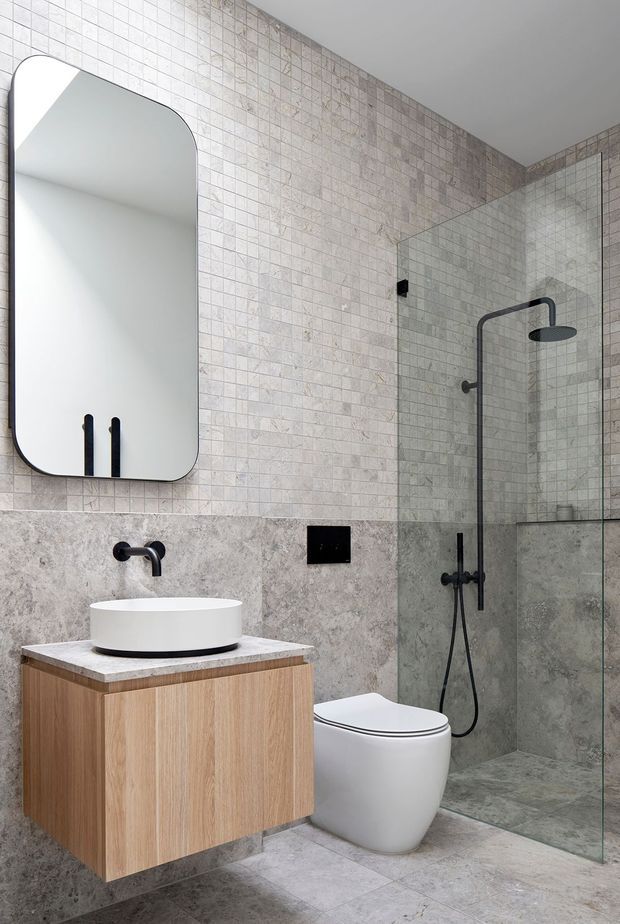 Contemporary bathroom with a walk-in glass shower, wall-mounted timber vanity, white vessel basin, wall-hung toilet, large rounded-corner mirror, and matte black tapware and shower fittings. The room is finished in pale grey stone-look mosaic wall tiles with larger matching stone-look floor and lower wall surfaces.