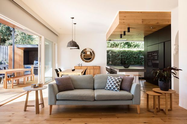 Open-plan contemporary living space with a grey upholstered sofa in the foreground, a timber dining table and sideboard behind it, and a dark minimalist kitchen at the rear. Full-height glazing opens toward a timber outdoor deck, while a warm timber-lined ceiling feature extends over the kitchen and dining area. Neutral finishes, natural timber flooring, pendant lighting, and indoor plants create a bright, warm, relaxed interior.