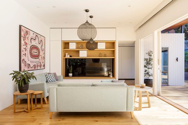 Bright contemporary living room with a pale upholstered sofa facing a built-in timber entertainment and shelving unit. The room features light timber flooring, white walls and ceiling, layered pendant lighting, large glazed sliding doors opening to an outdoor deck, and minimal furnishings with indoor plants and artwork.