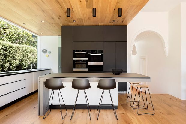 A bright contemporary kitchen with a large island and three dark stools, integrated appliances set within full-height charcoal cabinetry, and a warm timber-lined ceiling. White handleless perimeter cabinetry sits beneath expansive glazing overlooking greenery. The room also includes timber flooring, a sculptural arched opening, and additional bar seating.