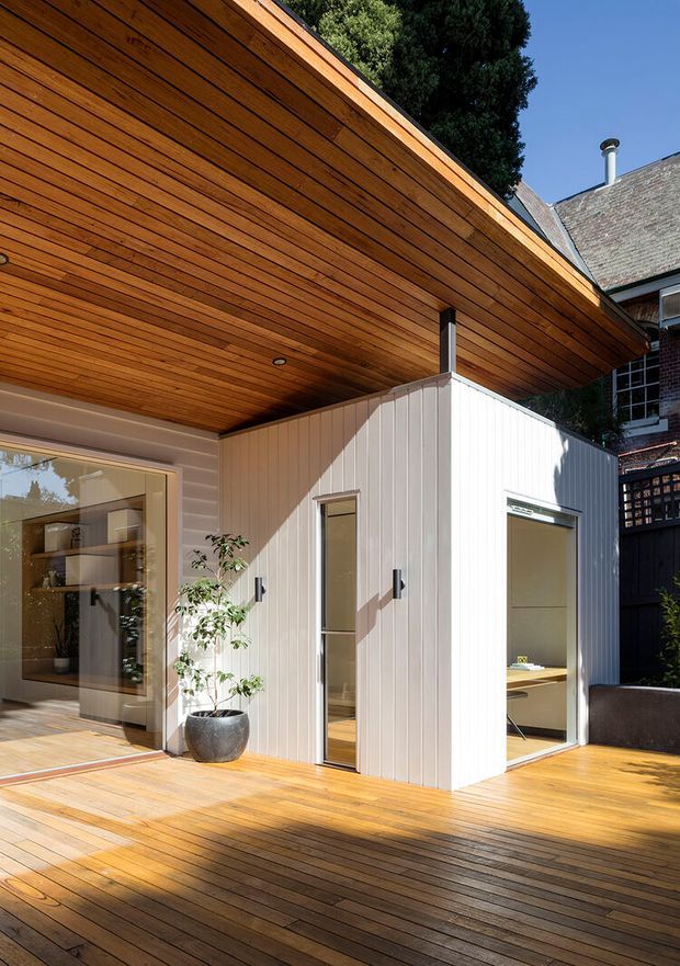 A bright architectural exterior view of a contemporary timber-clad extension opening onto a broad timber deck. The extension has a deep projecting roof with a warm timber-lined soffit, pale vertical wall cladding, large glazed sliding doors, narrow vertical windows, and recessed ceiling lighting. A compact interior workspace is visible through the openings, while an older brick-and-shingle building appears behind it, showing a modern addition integrated into an established residential setting.