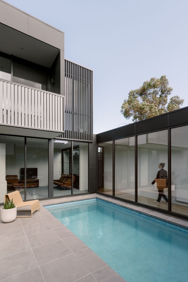 A contemporary multi-level home arranged around a compact rectangular swimming pool courtyard. The exterior features dark-framed floor-to-ceiling glazing, pale grey tiled paving, vertical metal screening, cantilevered upper-level balconies, and a flat roofline. Interior living areas are visible through the glazing, with a tree rising beyond the courtyard wall.