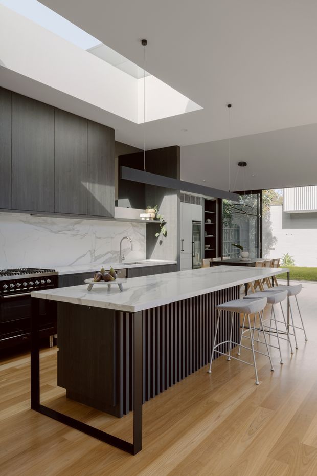 A contemporary open-plan kitchen with a large white marble-look island, dark timber cabinetry, integrated appliances, a ribbed island base, timber flooring, and a long linear pendant light. A roof skylight brings natural light into the space, with dining and garden views beyond.