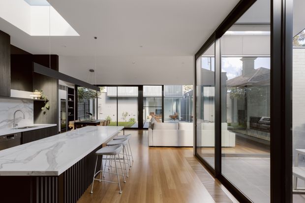 A bright, contemporary open-plan kitchen overlooking a glazed courtyard or outdoor area. The room features a long white stone island with integrated seating, dark timber cabinetry, warm timber flooring, extensive black-framed glazing, a skylight, and a white living or dining zone beyond.