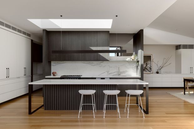 Contemporary open-plan kitchen with a large island and three stools, dark timber cabinetry, a dramatic white veined stone splashback, integrated appliances, and warm timber flooring. A skylight brightens the space, while adjoining white cabinetry and a dining area are visible in the background.