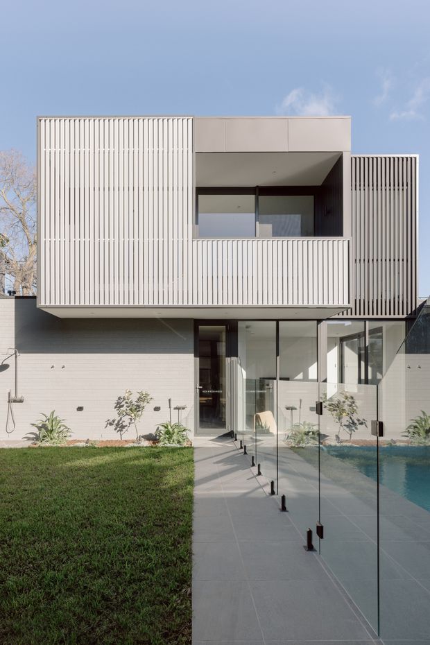 Front exterior view of a contemporary multi-level residence with crisp rectilinear forms, pale rendered walls, extensive vertical slatted screening, recessed glazing and a projecting upper volume. A paved path leads past frameless glass pool fencing toward the entry, with a swimming pool visible at right and a small landscaped lawn and garden along the facade.