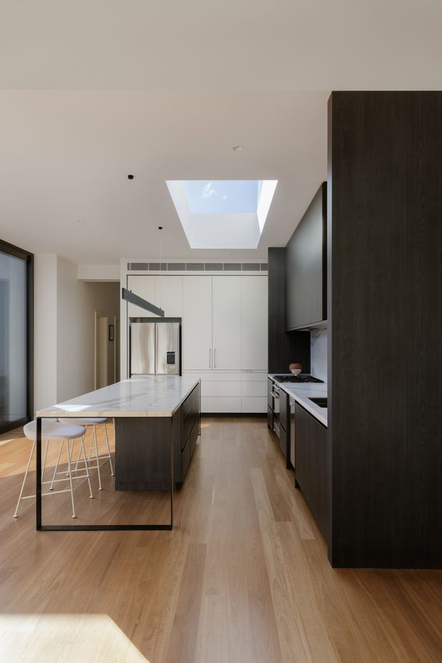 A contemporary galley-style kitchen viewed from the living area, featuring a large white stone island with an integrated dark timber base and black-framed glass end panel, white full-height cabinetry, dark wood cabinetry along the right wall, timber flooring, stainless-steel appliances, and a large skylight providing natural light. Minimal furnishings and clean linear detailing create a refined, bright interior.