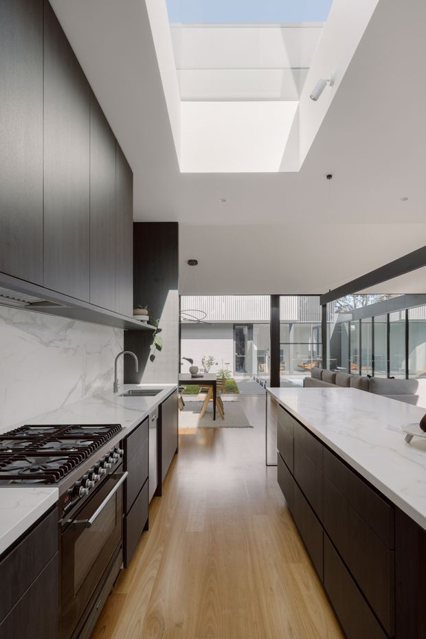 A contemporary galley-style kitchen opening into a bright open-plan living and dining area. The kitchen features dark timber-look cabinetry, a large white marble-look island and benchtops, a pale timber floor, integrated stainless-steel appliances, and a dramatic rectangular skylight. Full-height glazing connects the interior to a courtyard-like outdoor space.
