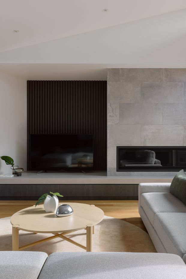 Contemporary living room featuring a dark vertically ribbed timber TV wall, pale large-format stone or porcelain fireplace surround, integrated black fireplace, and low dark built-in cabinetry. A light grey sectional sofa, pale round timber coffee table, neutral rug, and restrained decor create a refined, warm minimalist interior.