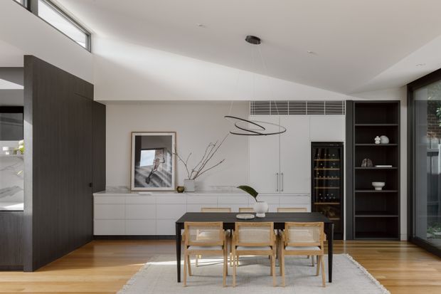 A refined open-plan dining and kitchen area with a timber dining table and woven-back chairs positioned on a pale rug. The room features light timber flooring, white built-in cabinetry, a dark timber storage wall, integrated wine storage, open shelving, large glazing, and a sculptural linear pendant light. The palette is warm neutral with strong dark-wood cabinetry accents.