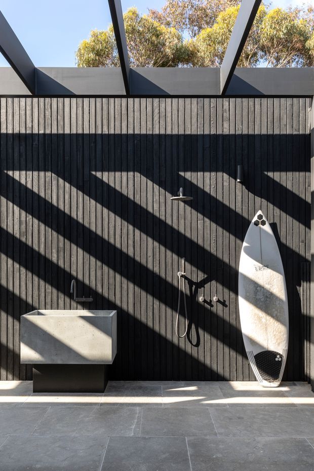A contemporary outdoor shower area with a freestanding concrete basin against a dark vertical timber-clad wall. A surfboard leans beside exposed shower fixtures, while strong geometric shadows fall across the wall and roof beams. Large-format grey paving, a partially open overhead structure, and eucalyptus trees visible above the wall create a modern coastal outdoor setting.