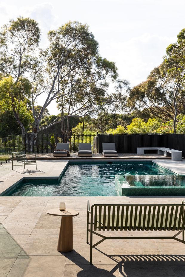 A landscaped outdoor pool terrace surrounded by mature trees and dense greenery. The geometric swimming pool includes a raised spa or water feature, with pale stone paving, dark metal fencing, built-in outdoor seating, sun loungers, and a contemporary slatted bench and side table in the foreground.