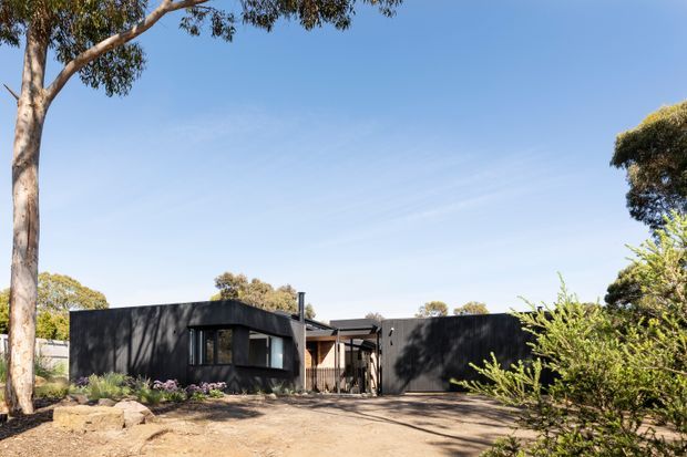 Single-storey contemporary dark-clad house set within a landscaped, dry garden. The low, horizontally composed building has dark walls, broad glazing, a sheltered central entry, and a flat or very low-pitched roof, surrounded by mature trees and shrubs.