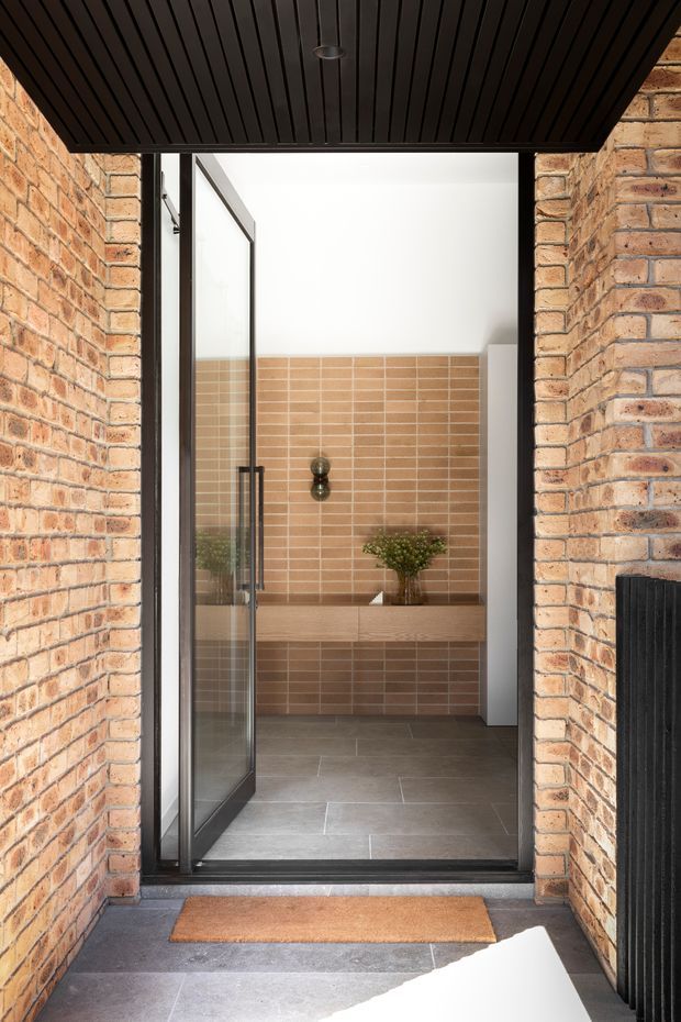 View through a black-framed glass door into a refined bathroom or powder room. The entrance is framed by exposed warm brick walls and a dark slatted ceiling. Inside, a tan rectangular-tiled feature wall sits above a floating timber-toned vanity with an integrated basin, black wall-mounted tapware, and a small plant. Large grey floor tiles and black architectural framing create a warm contemporary industrial character.