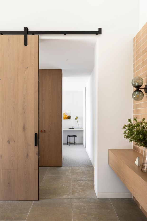 A bright, refined residential hallway viewed through a partially open, large light-oak barn-style sliding door. The space features warm timber cabinetry and a floating timber console, large taupe stone-look floor tiles, white walls, a black metal door track and hardware, and an exposed light brick feature wall with globe wall lighting. A small desk and stool are visible at the end of the corridor.