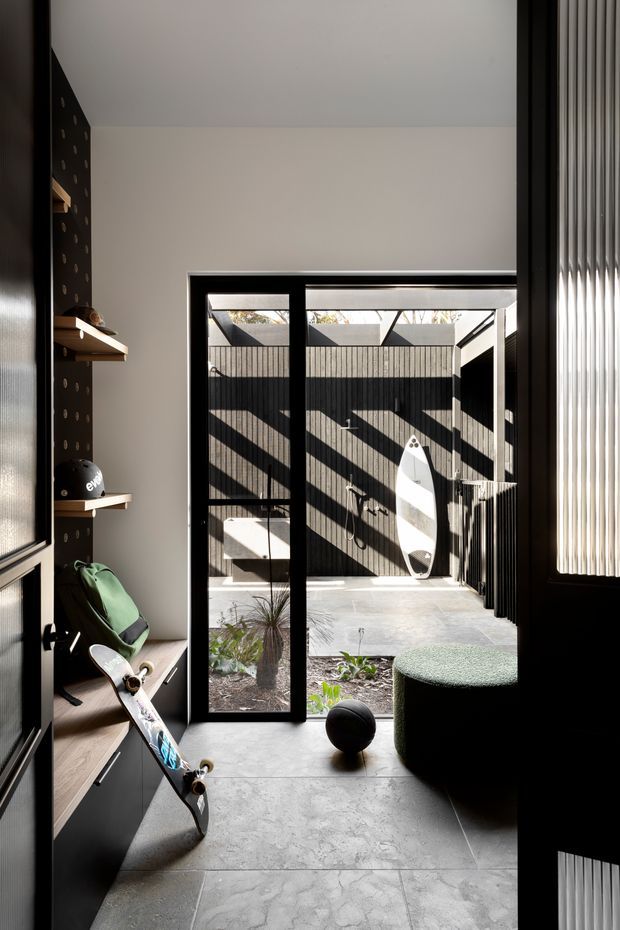 A contemporary indoor-outdoor transition space with dark built-in cabinetry and shelving, a skateboard, sports equipment, and a green upholstered ottoman. Large black-framed glazed doors open to a compact paved courtyard with a surfboard mounted against a dark slatted fence, while strong sunlight creates geometric shadows across the space. A vertical ribbed glass or screen element frames the right side.