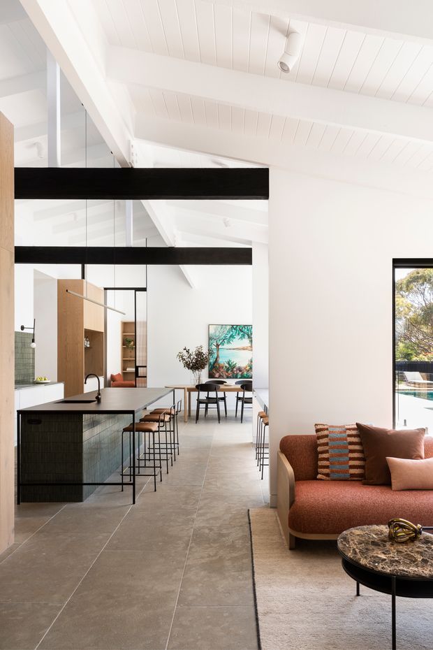 Bright open-plan residential interior combining a kitchen, dining area and living space. The room features white painted timber-lined vaulted ceilings with exposed beams, large internal glazing, a dark island with integrated seating, pale stone or concrete-look floor tiles, warm timber joinery, a rust-toned upholstered sofa, and a marble-topped round coffee table. The composition is airy and contemporary with strong black structural accents.