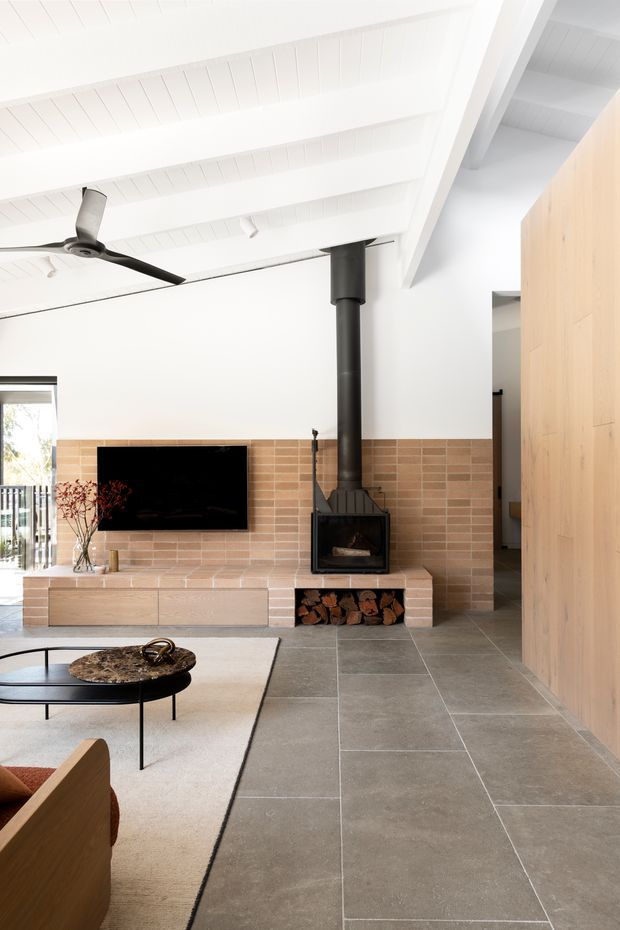 Bright, open-plan living room with a white pitched, exposed-beam ceiling, black ceiling fan, large grey stone-look tiled floor, and a warm timber feature wall. A brick-lined fireplace with black wood-burning stove and integrated firewood storage forms the focal point, alongside a wall-mounted television, low timber joinery, coffee table and neutral rug. Glazing at left opens toward an exterior balcony or deck.