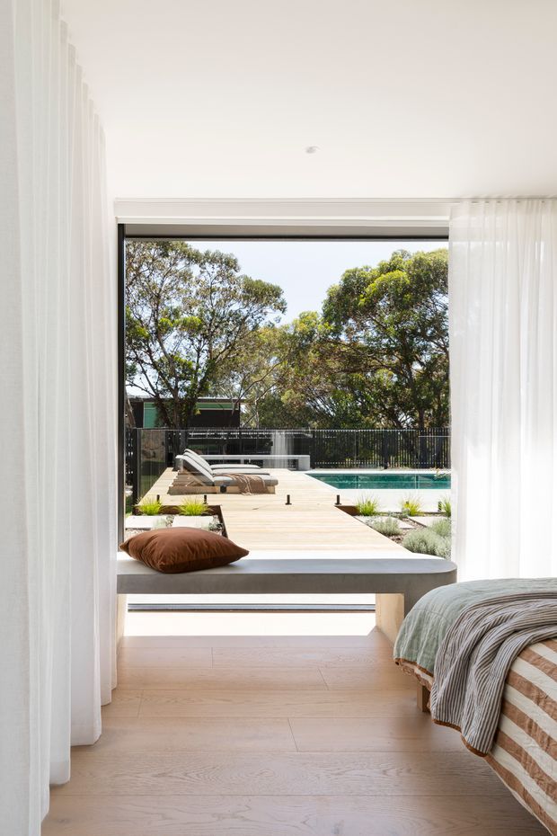 Bright, serene bedroom opening directly onto a landscaped timber pool deck. The room has pale timber flooring, white sheer curtains, a broad floor-to-ceiling opening, and a partially visible bed with muted green-grey bedding. Outside is a built-in window seat or daybed, timber decking, loungers, planting, fencing, and a swimming pool.