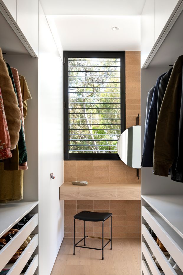 A bright, compact walk-in wardrobe with open white built-in clothing storage and drawers on both sides. A timber-lined bench sits beneath a large black-framed horizontal-louvered window, with a round mirror and black stool completing the dressing area. The room has pale timber flooring, warm tan brick or tile wall cladding, and a clean contemporary fit-out.