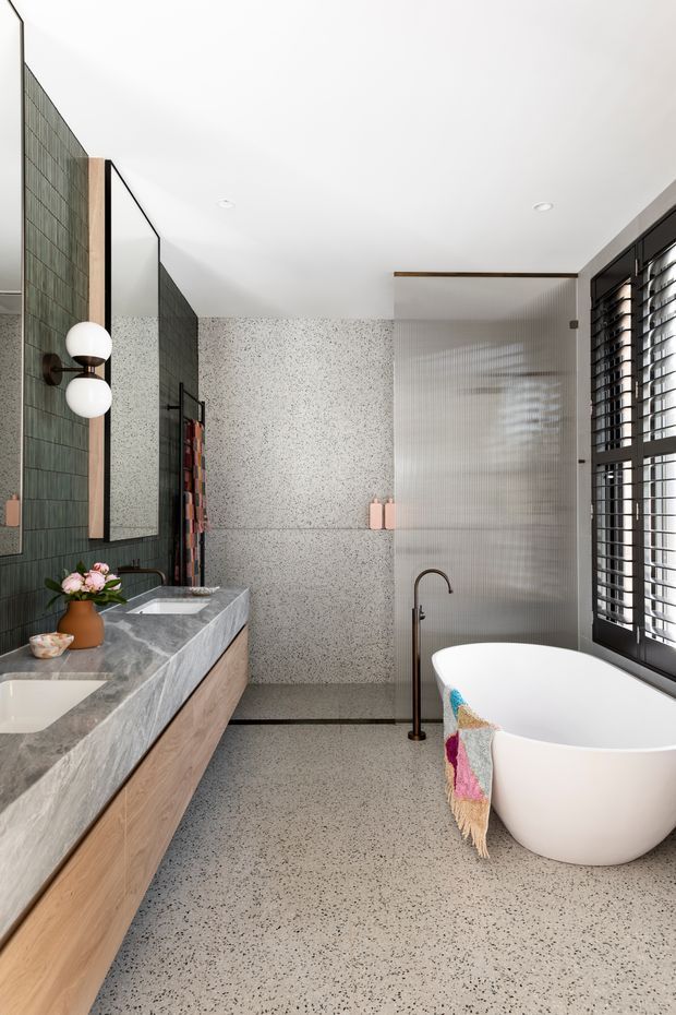A bright, refined contemporary bathroom featuring a freestanding white bathtub, wall-mounted basin vanity, large grey stone-look benchtop, timber cabinetry, terrazzo-style wall and floor surfaces, dark green vertical wall tiles, a full-height mirror, ribbed translucent glass screen, and black-framed louvred window shutters. A sculptural double globe wall light and colourful bath textile add detail.