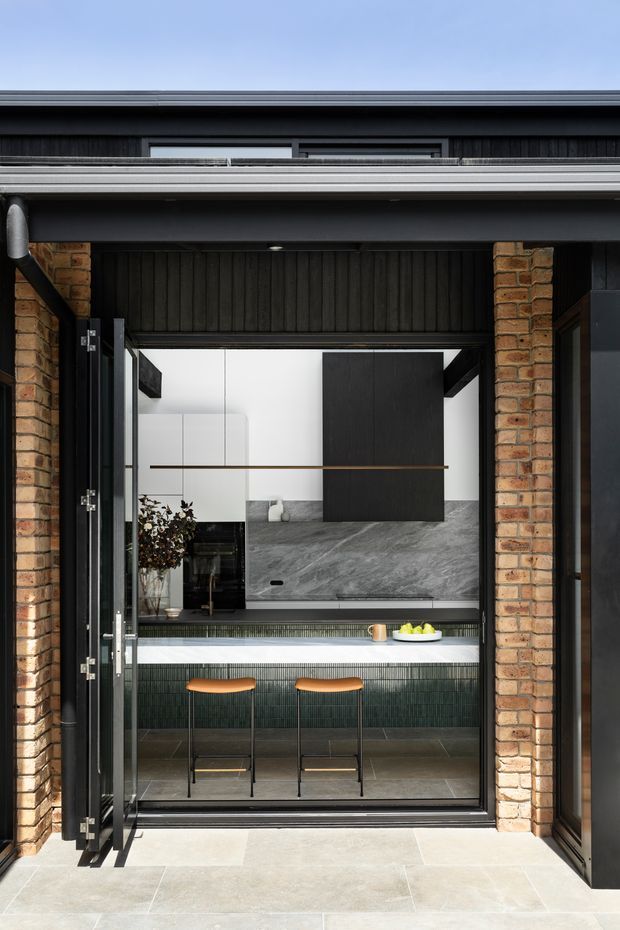 A contemporary kitchen and breakfast bar viewed through a large open doorway from an exterior terrace or courtyard. The design combines black vertical timber cladding and cabinetry, white upper cabinetry, a dramatic grey stone splashback and benchtop, dark green vertically textured tiles on the island front, warm timber bar stools, exposed brick piers, and dark metal framing. The composition highlights the indoor-outdoor connection and strong contrast between light stone paving and dark architectural finishes.