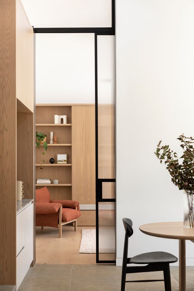 A refined contemporary interior viewed from a dining area toward a living room. The space features warm timber built-in shelving and wall panelling, pale wood flooring, a black-framed ribbed glass partition, a round timber dining table with black dining chair, and a rust-coloured upholstered armchair. Neutral white walls and natural greenery create a calm, warm atmosphere.