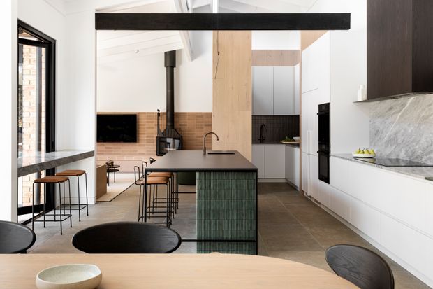 Open-plan contemporary kitchen and dining area with a dark stone island, green vertical-tile island front, white cabinetry, timber accents, a large marble-look splashback, and an adjoining living space with a freestanding wood-burning stove. Bar stools and a dining table occupy the foreground, while exposed structural and ceiling elements add architectural character.