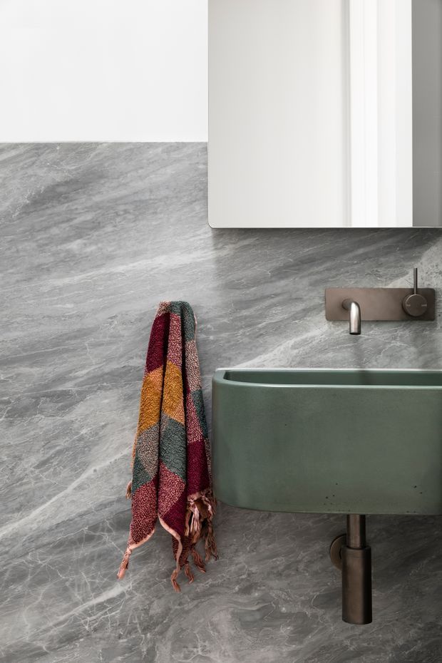 Close-up of a contemporary bathroom featuring a wall-mounted muted green basin with exposed dark metal plumbing, a brushed metal wall-mounted faucet, a large grey veined stone-effect wall finish, and a white mirrored or wall cabinet above. A multicoloured textured towel hangs beside the basin.