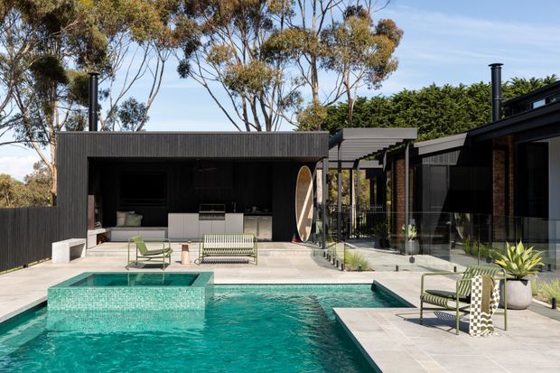 Contemporary single-storey home with a dark vertical-clad pavilion framing a landscaped pool courtyard. The outdoor area includes a turquoise swimming pool with raised spa, pale stone paving, an open-air kitchen and barbecue, glass walls, black-framed glazing, and outdoor lounge furniture. Mature trees and hedging surround the private courtyard.