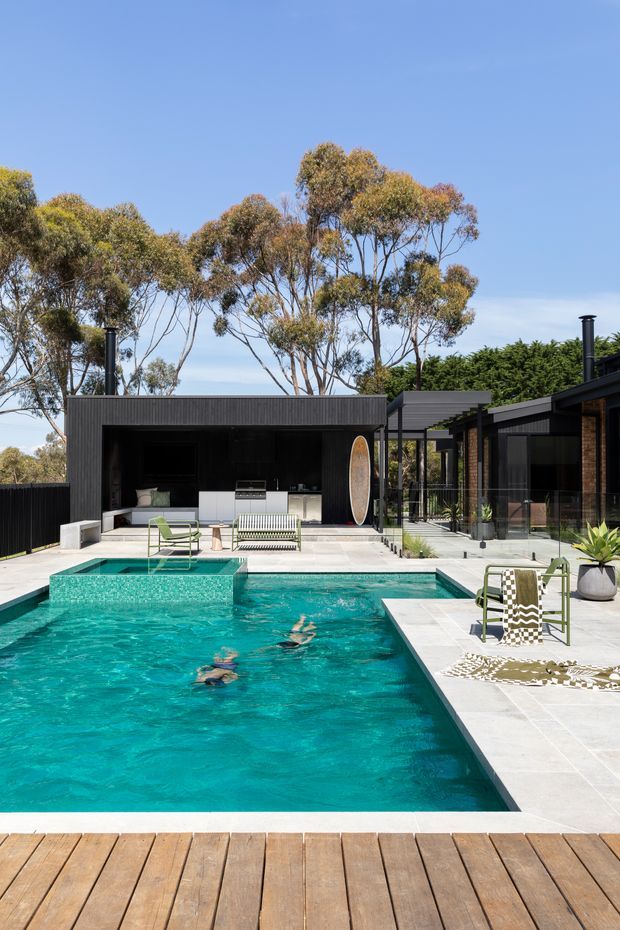 A contemporary single-storey home arranged around a bright turquoise swimming pool. The outdoor area features pale tiled paving, a timber pool deck, black-clad pavilion and house volumes, an outdoor kitchen, glass fencing, lounge chairs, and mature eucalyptus trees beyond.