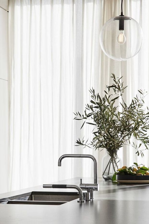 Bright, minimalist kitchen detail featuring a dark grey island benchtop with an integrated double sink and two chrome gooseneck taps. Sheer white curtains provide a softly lit backdrop, complemented by a clear glass globe pendant light and a vase of leafy branches.