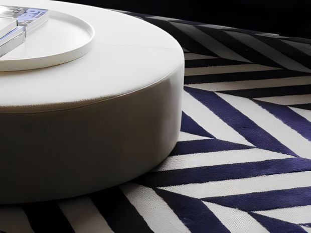 Close-up interior detail of a low, rounded white upholstered coffee table or ottoman positioned on a bold geometric rug. The rug features irregular black, white and deep blue angular stripes, creating a graphic contemporary look. A white tray with stacked magazines sits on the tabletop.