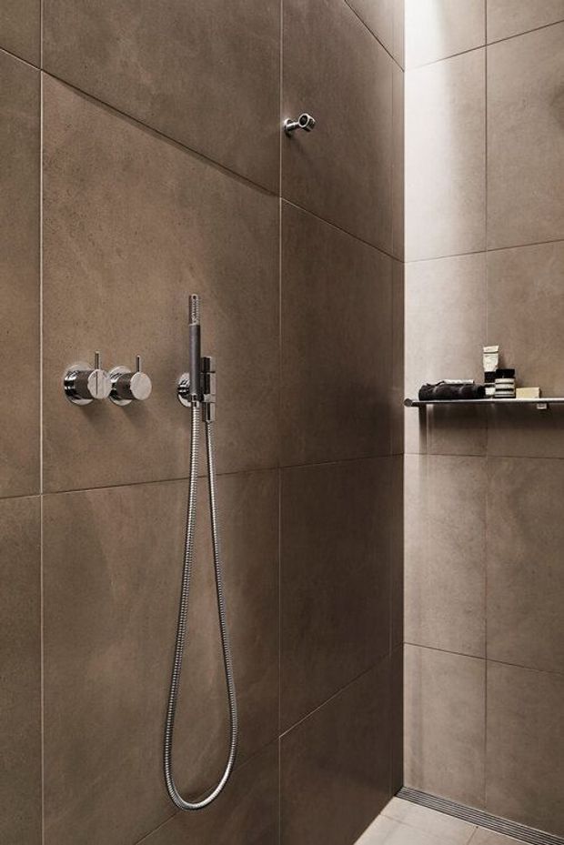 Close-up view of a contemporary walk-in shower with large-format taupe-brown wall tiles, chrome shower controls and handheld shower hose, a recessed corner shelf with toiletries, and a linear floor drain. The space has a clean, minimalist, refined appearance.