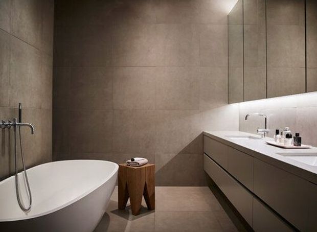 Contemporary bathroom with a freestanding white bathtub, wall-mounted shower hardware, large-format taupe stone-look tiles, a floating dark vanity with integrated basins, and mirrored upper cabinetry. Concealed linear lighting beneath the cabinets creates a warm, refined atmosphere.